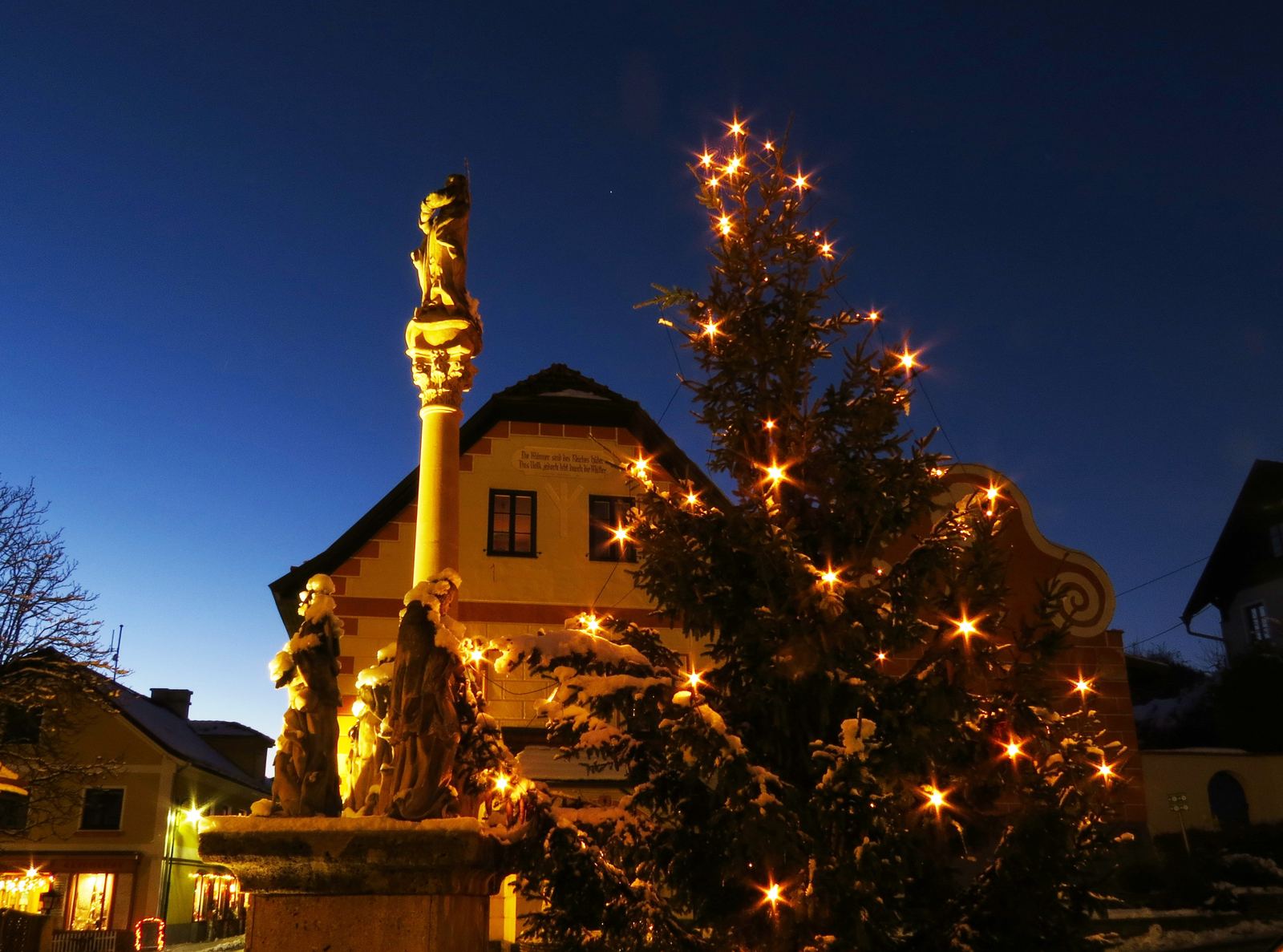 Beleuchteter Christbaum und Mariensäule am verschneiten Marktplatz am Abend